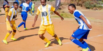 Young soccer players in yellow and blue uniforms competing on a sandy field, with spectators and a 10bet.co.za banner in the background.