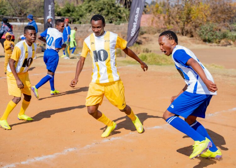 Young soccer players in yellow and blue uniforms competing on a sandy field, with spectators and a 10bet.co.za banner in the background.